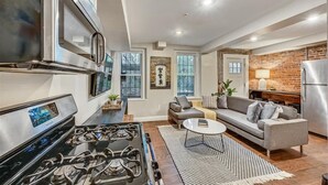 A modern kitchen with a stainless steel range hood, a built-in oven, and a gas stove. There is a gray sofa and a small round table in the living area. - Clean contemporary escape (Boston)
