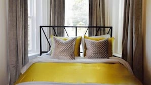 Una cama con estructura metálica, cortinas grises y una colcha amarilla.