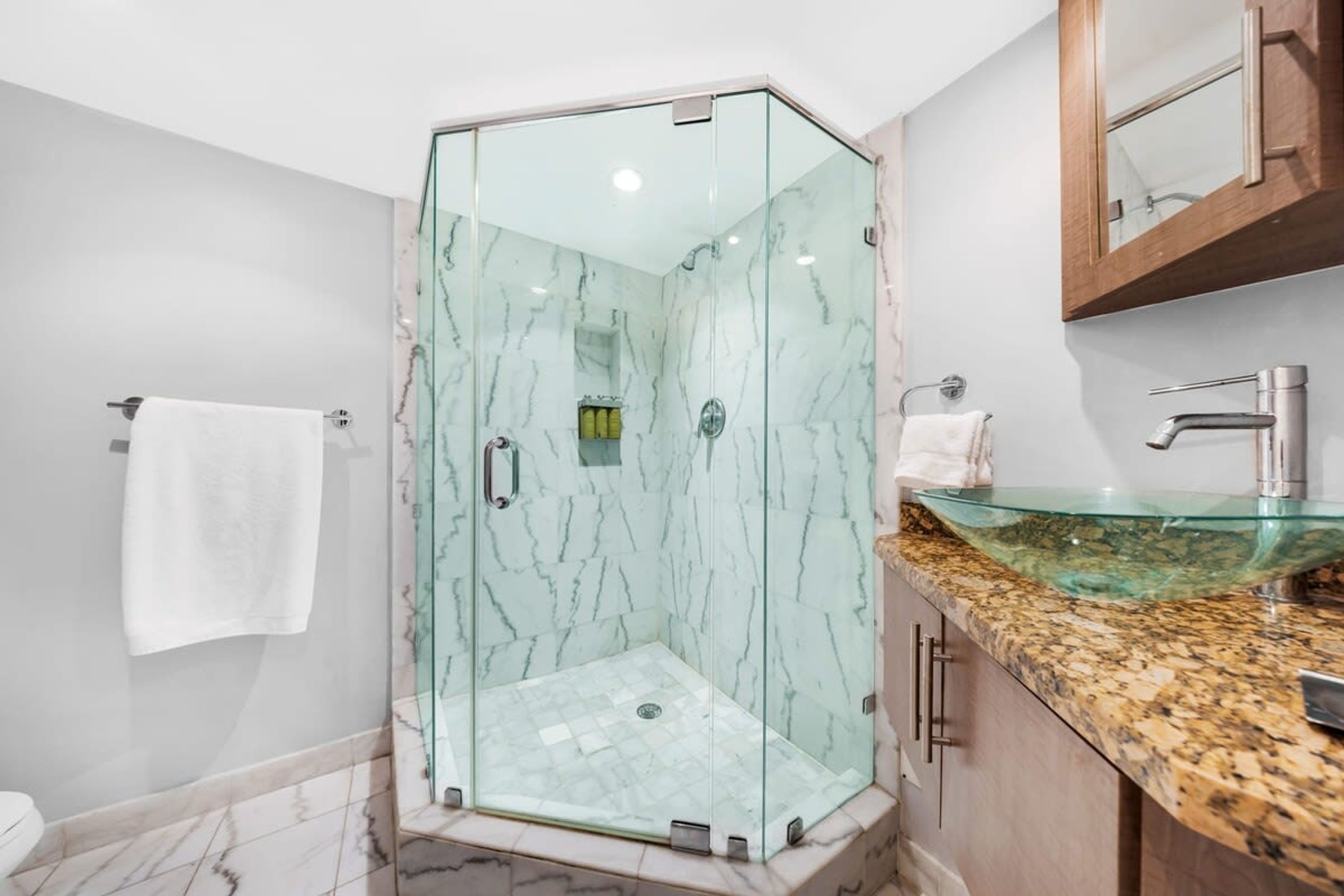 A modern bathroom with a glass-enclosed shower, a marble-tiled floor, and a granite countertop with a glass sink.