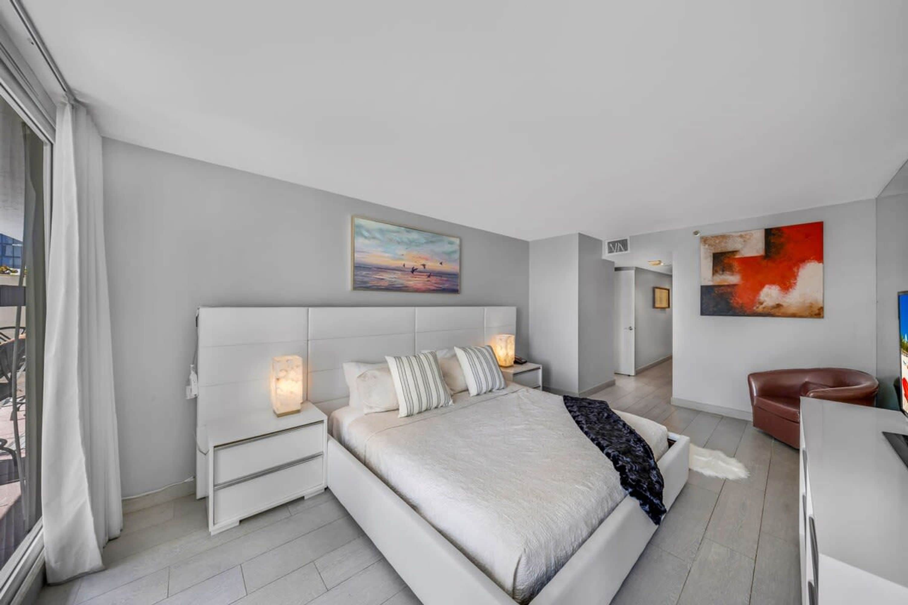 A modern bedroom with a large bed, bedside tables, a desk, and a chair. There are two paintings on the walls and a view of the outdoors through a window.