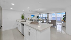 A modern kitchen with a large island, white cabinetry, and a view of the city through large windows. - The Grand Wonder Biscayne (Miami)