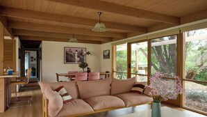 A modern living room with a wooden ceiling, a sofa, a dining area, and large windows with a view of the outdoors.