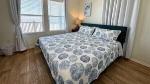Un dormitorio con cama, ventana con persianas, mesita de noche con lámpara y un cuadro en la pared.
