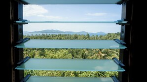 Uitzicht door een raam met meerdere horizontale zonwering, op een weelderig groen landschap met bomen en verre bergen.