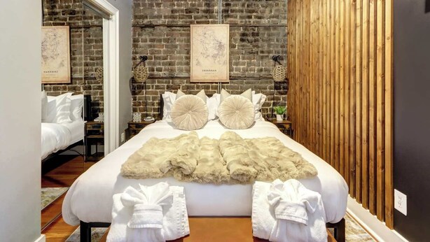 Une chambre avec un mur en briques, un mur avec des lambris en bois et un lit recouvert de draps blancs et agrémenté d’oreillers décoratifs.