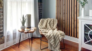 Une pièce chaleureuse avec un plancher en bois, un fauteuil agrémenté d’une couverture à motifs, une petite table ornée d’une plante et d’un vase décoratif, et une bibliothèque remplie de livres et d’un objet décoratif.