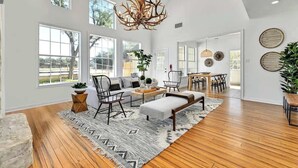 Un salon spacieux avec un plafond haut, de grandes fenêtres, un plancher en bois, un canapé, une table basse et un lustre.