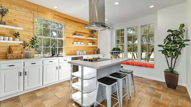 Une cuisine moderne avec des meubles blancs, un îlot central doté de tabourets, un lambris en bois et de grandes fenêtres.