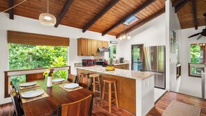 Une cuisine moderne avec des poutres en bois, un grand îlot central doté de tabourets et une vue sur une végétation luxuriante.