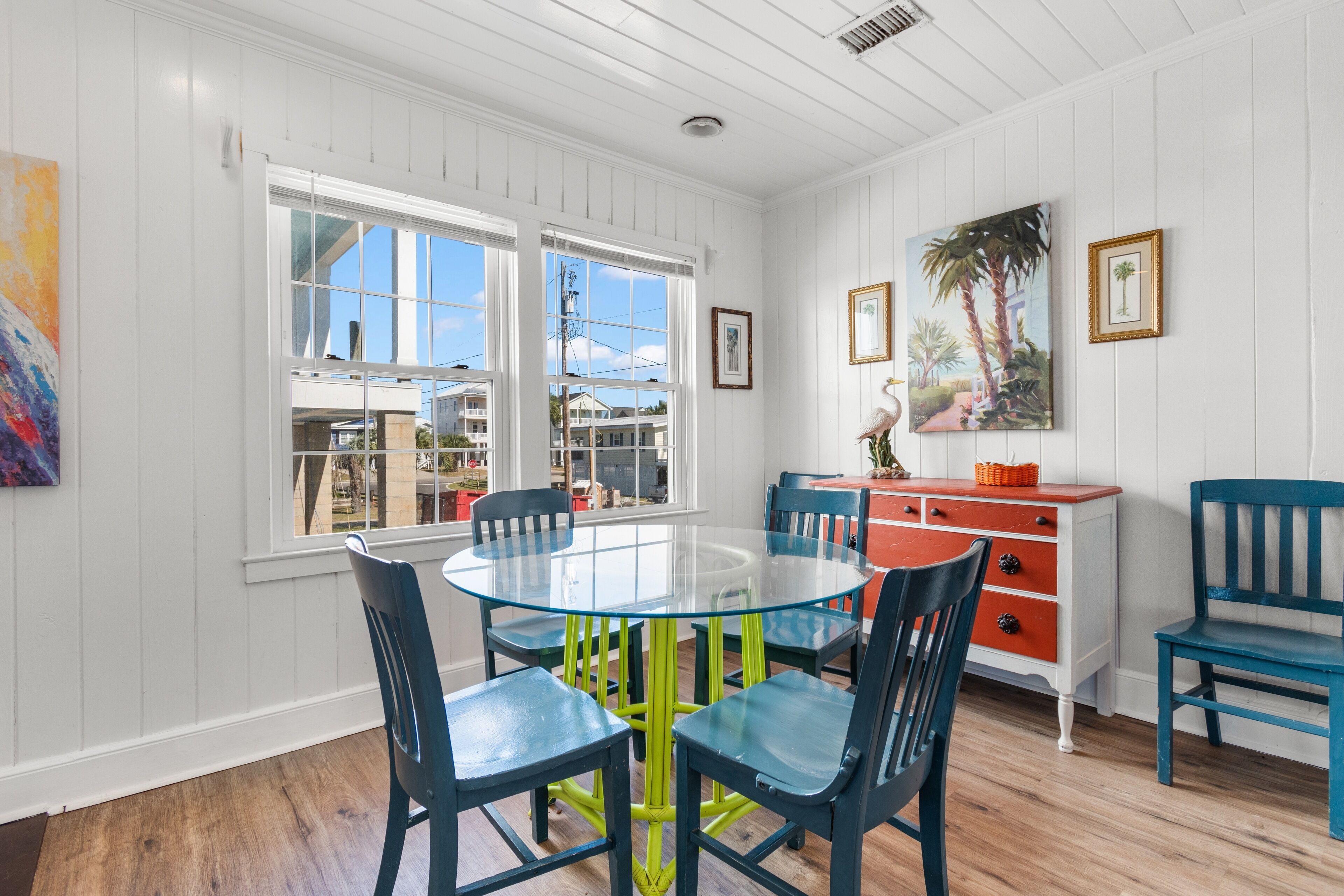 A dining area with a glass table surrounded by blue chairs, a red and white cabinet, and a painting of palm trees.