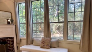 Ein gemütliches Zimmer mit einer weißen Bank vor einem großen Fenster, einem Backsteinofen und Blick auf einen Wald.