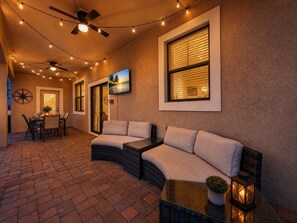 An outdoor patio area with a curved sofa, dining table, chairs, string lights, ceiling fans, and a large window.