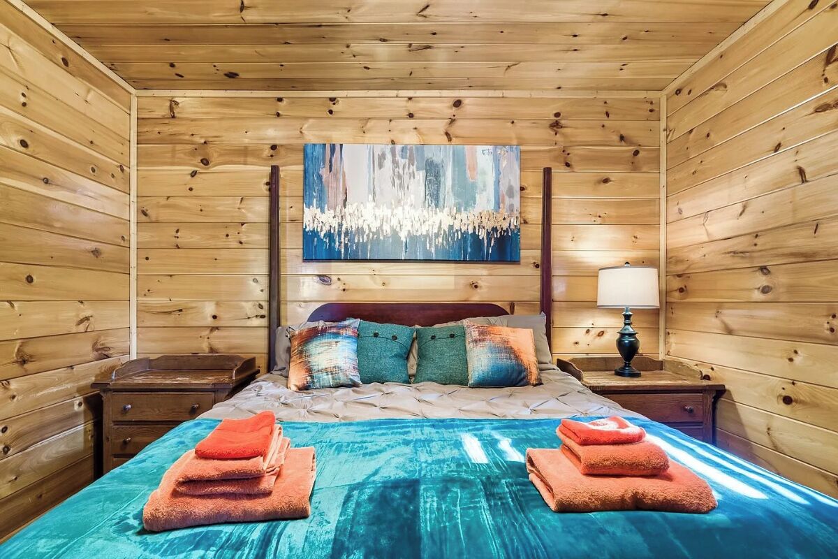 A wooden-paneled room with a bed, a painting above the bed, and a nightstand with a lamp.