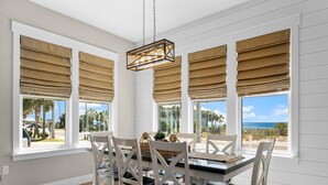 Et spiseområde med et bord dækket til seks personer, omgivet af hvide stole, store vinduer med beige persienner og en hængende lampe.