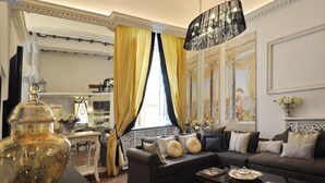 A living room with a sofa, a coffee table, and a chandelier. There is a window with yellow curtains and a painting on the wall.