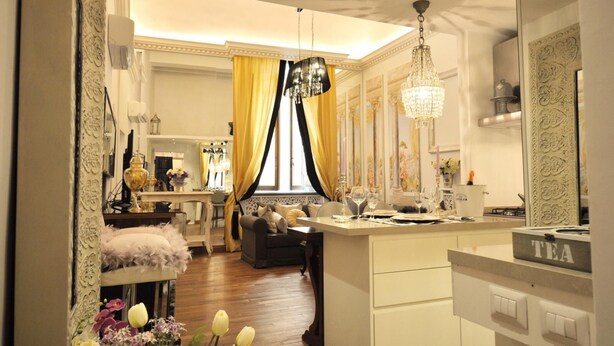 A well-lit, elegant dining area with a chandelier, a kitchen area with a tea sign, and a view of the dining room through a window with yellow curtains.