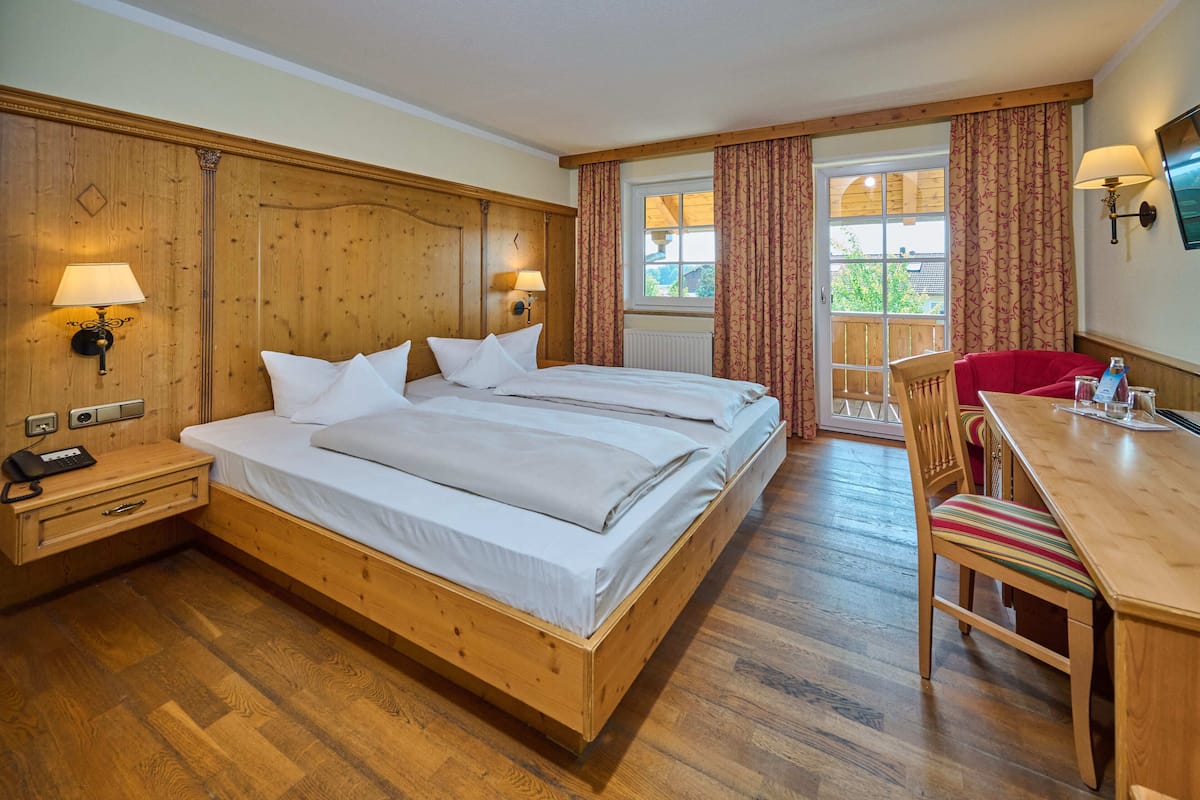 Une chambre d’hôtel avec un lit en bois, deux tables de chevet avec des lampes, un bureau avec une chaise et une vue sur l’extérieur par une fenêtre.