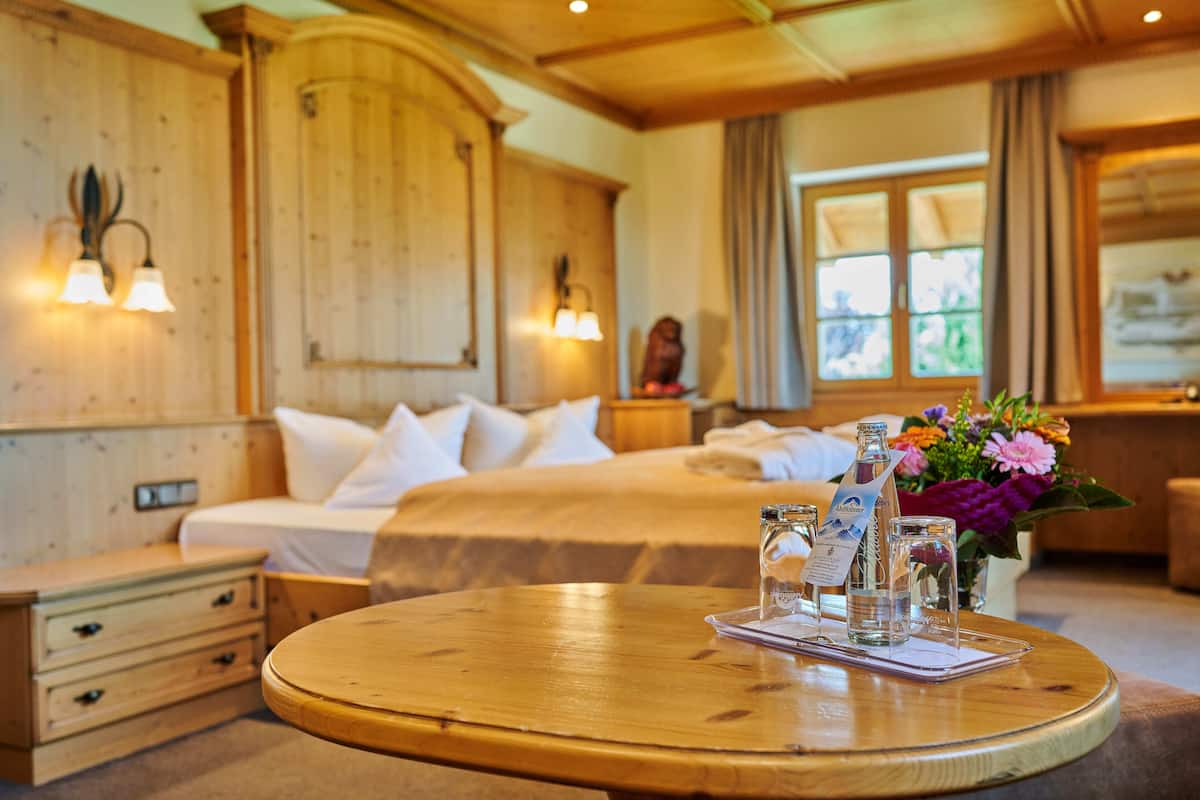 Une chambre d’hôtel avec un intérieur en bois, un lit avec du linge de maison blanc, une table ronde en bois sur laquelle est posé un menu, et un vase rempli de fleurs.