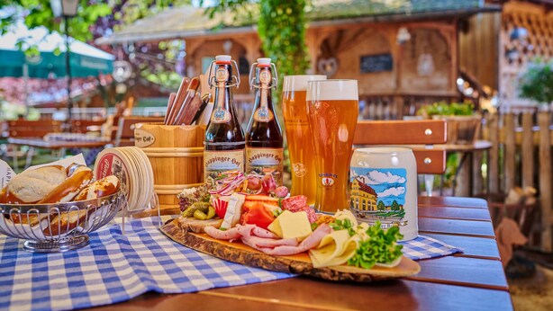 Une table en bois recouverte d’une nappe à carreaux, une variété de fromages, de charcuteries et de cornichons, ainsi que des bouteilles et des verres de bière.