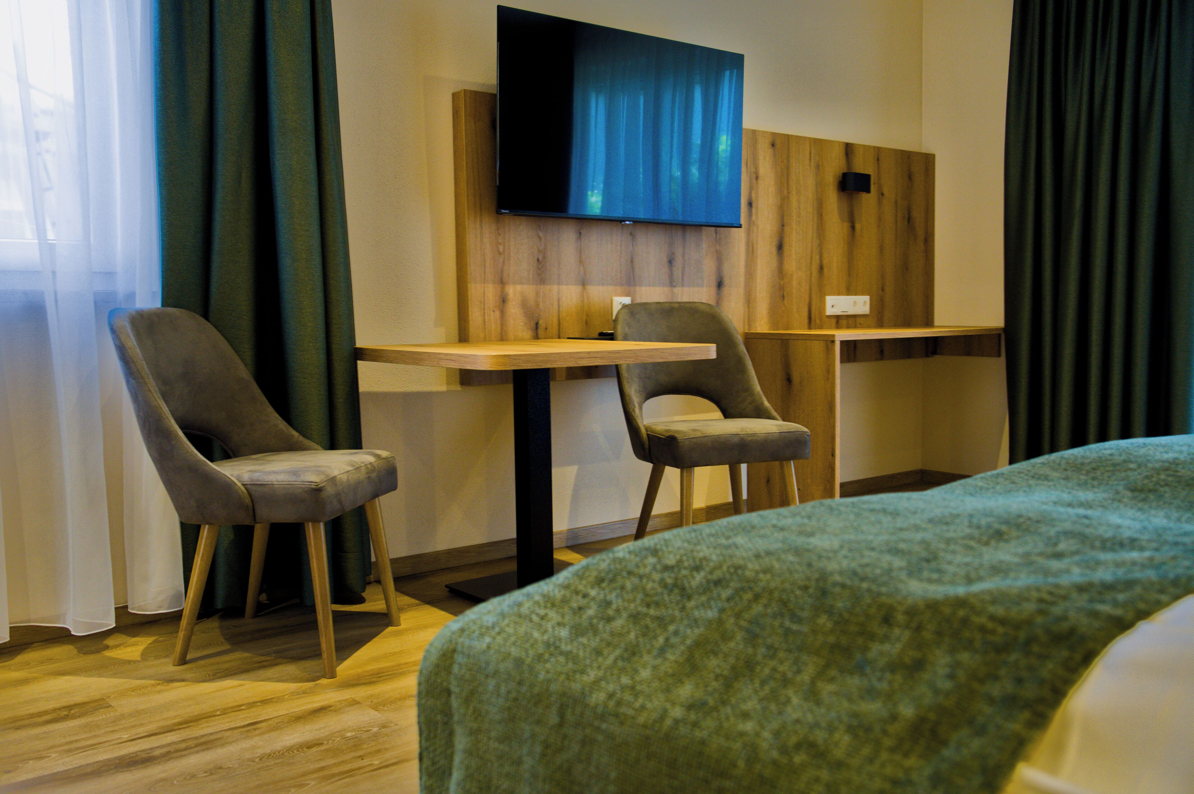 A hotel room with a bed, two chairs, a small table, a TV mounted on the wall, and wooden paneling.