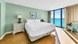 A hotel room with a large bed, a nightstand with a lamp, a view of the ocean through large windows, and a wicker chair by the window. - ⛵Gulf Front with Fab Amenities-Edgewater 3-1104⛵ (Panama City Beach)