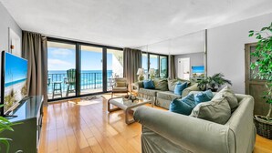 A spacious living room with a large sofa, a coffee table, and a TV. The room has large windows offering a view of the beach. - ⛵Gulf Front with Fab Amenities-Edgewater 3-1104⛵ (Panama City Beach)