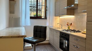 A modern kitchen with a gas stove, oven, and stainless steel sink. There is a window with a view of a brick building and a chair in the corner. - Il nido dell'Upupa (Montepulciano)