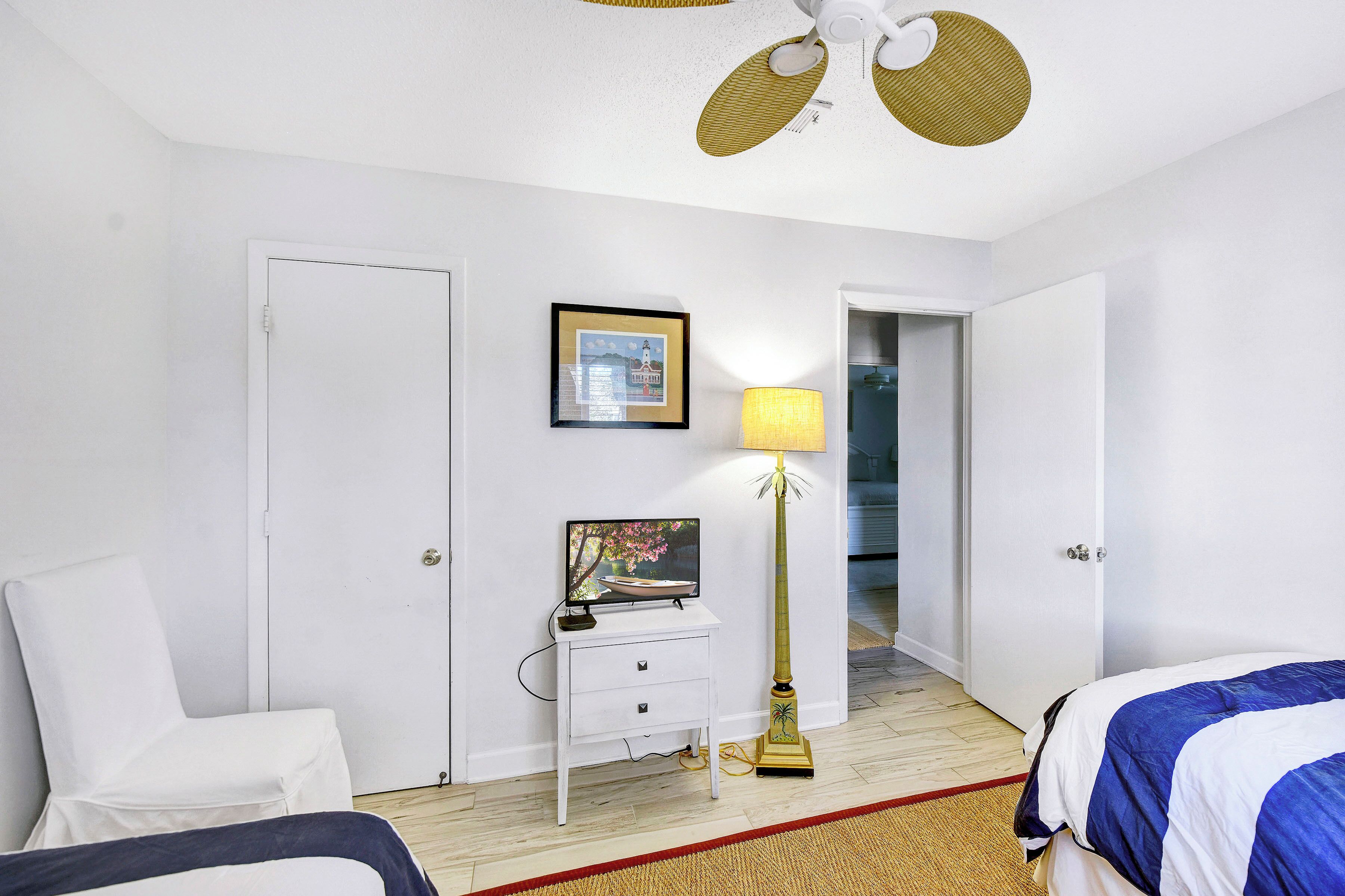 A hotel room with a white bed, a white chair, a bedside table with a lamp, a framed picture on the wall, and a ceiling fan.