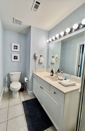 A bathroom with a toilet, sink, mirror with lights, and two hair dryers on the wall. - Perfect Just For You! Excellent spot right on the beach. (Panama City Beach)