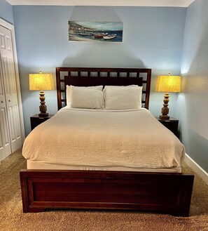 A bed with a wooden headboard, two bedside lamps, and a painting of a coastal scene on the wall. - Perfect Just For You! Excellent spot right on the beach. (Panama City Beach)