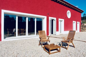 Un bâtiment rouge avec de grandes fenêtres et une terrasse en gravier agrémentée de chaises et d’une table en bois.