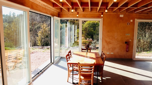Une pièce spacieuse avec une grande table à manger en bois et des chaises, de grandes fenêtres donnant sur l’extérieur, et un plafond avec des poutres apparentes et des luminaires suspendus.