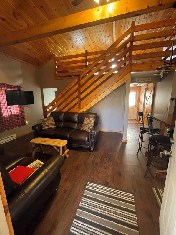 Un salon chaleureux avec un canapé en cuir, une table basse en bois et un tapis à rayures. Un escalier en bois mène à l’étage supérieur.