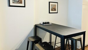 A small, modern kitchenette with a dark countertop, two stools, and framed coffee-themed artwork on the wall. - 7th Floor 1-Bedroom Condo with Parking (Calgary)