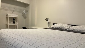 A bedroom with a bed, a desk, a chair, a shelf with towels, and a lamp. - 7th Floor 1-Bedroom Condo with Parking (Calgary)