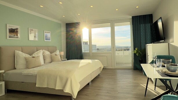 Ein Hotelzimmer mit Bett, Esstisch mit Stühlen, Fernseher und Blick auf die Stadt durch große Fenster.