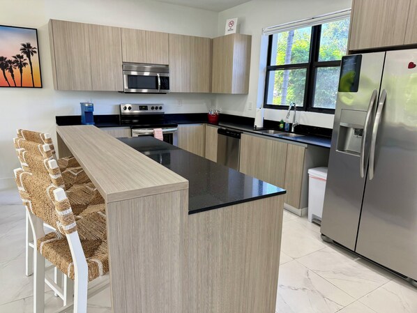 Una cocina moderna con armarios de madera clara, encimera negra, electrodomésticos de acero inoxidable y una ventana que proporciona luz natural.
