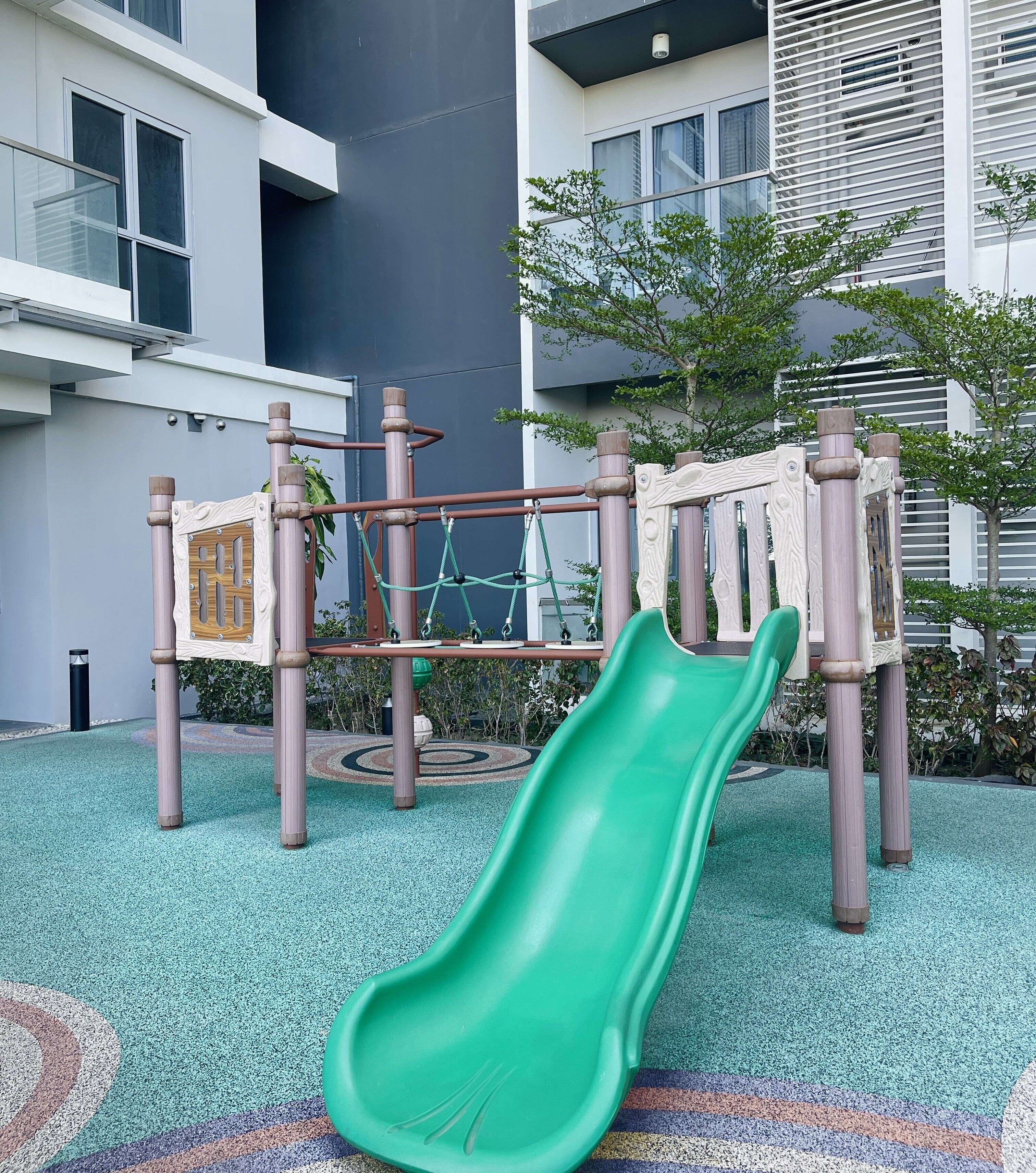 A playground with a green slide and climbing structures, surrounded by a building with large windows and a patterned floor.