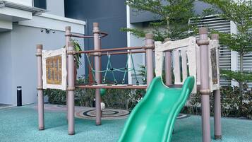 A playground with a green slide and climbing structures, surrounded by a building with large windows and a patterned floor.