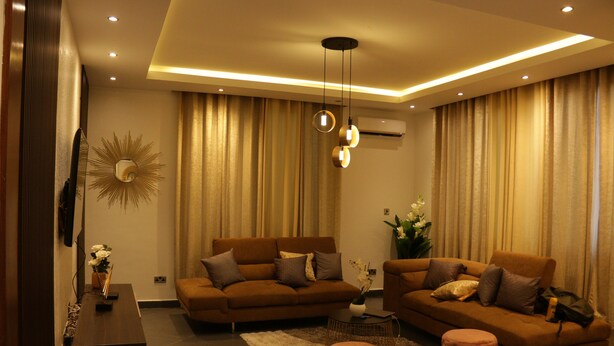Un salon moderne avec un ensemble de canapés marron, une table basse et deux poufs. Un miroir décoratif en forme de soleil orne le mur, et un luminaire suspendu éclaire la pièce.