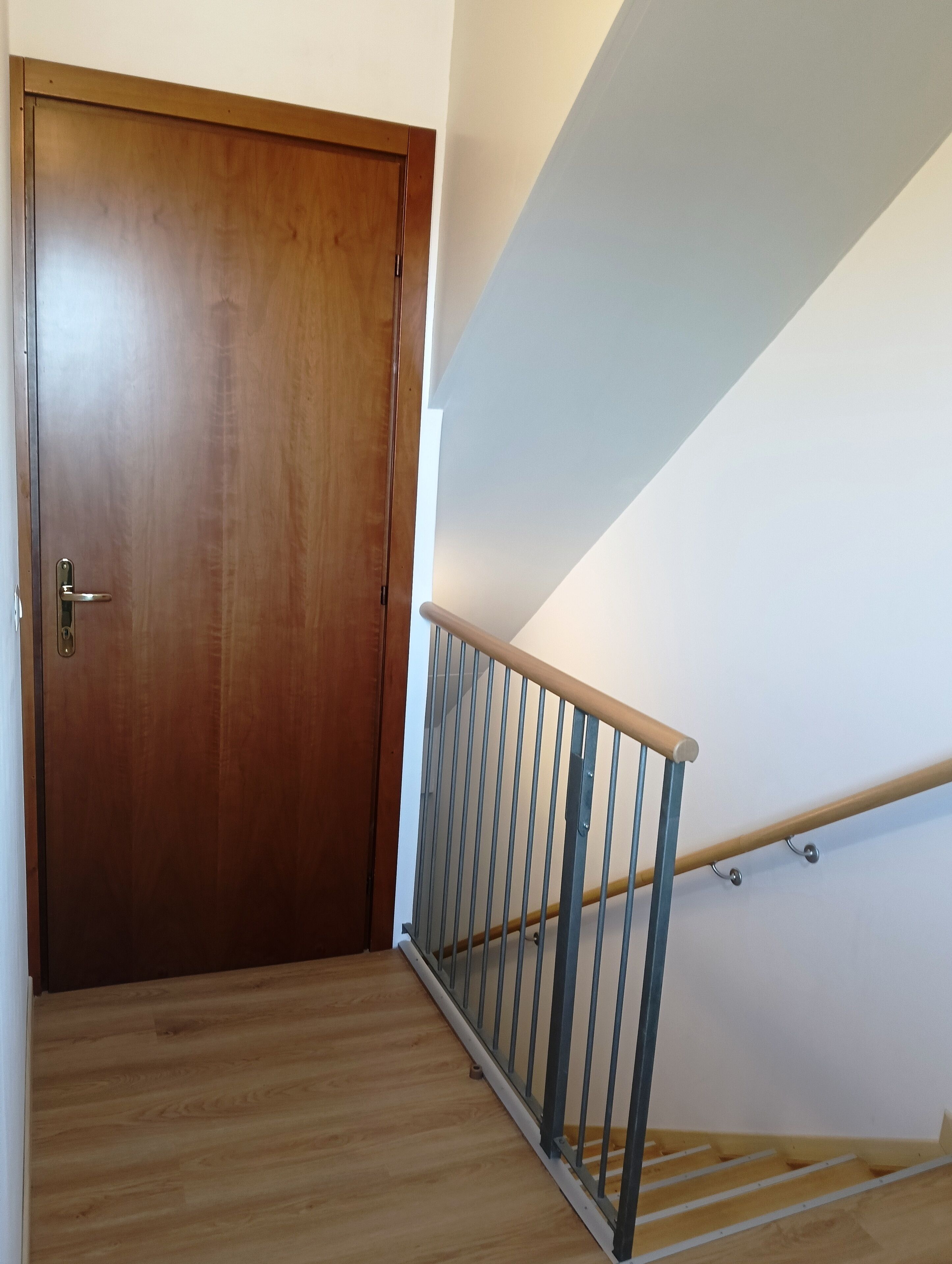 Una porta in legno con maniglia metallica, una scala in legno con ringhiera metallica e una parete bianca.