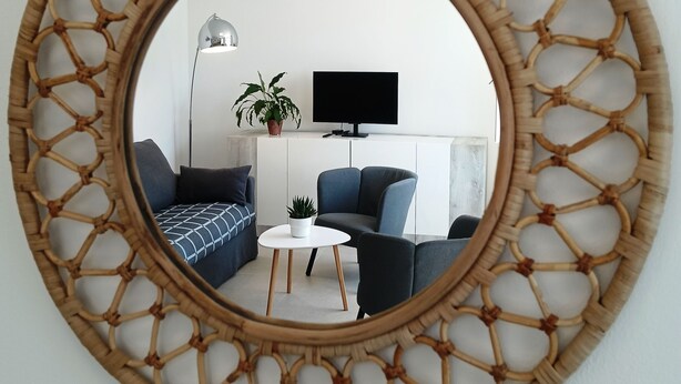 Un miroir rond doté d’un cadre tressé, qui reflète un salon avec un canapé, une table basse, une plante et un téléviseur.