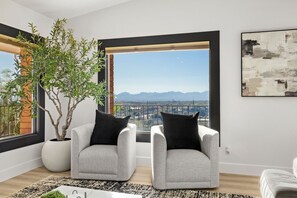 A modern living room with two grey armchairs, a large potted plant, and a view of mountains through a large window.