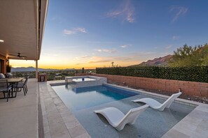Une piscine avec des chaises longues, un espace patio doté d’une table et de chaises, et une vue sur les montagnes au coucher du soleil.