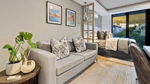 A modern living room with a grey sofa, a round side table with a plant, and a large window with a view of buildings and greenery. - Luxury Living @ Station House | No loadshedding (Cape Town)