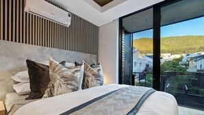 A bedroom with a large bed, a modern headboard, and a view of buildings through a large window. - Luxury Living @ Station House | No loadshedding (Cape Town)