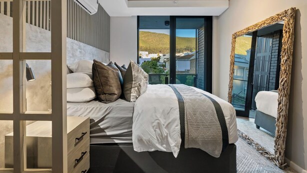 A modern bedroom with a large bed, a wooden dresser, and a view of a hillside through a glass door. - Luxury Living @ Station House | No loadshedding (Cape Town)