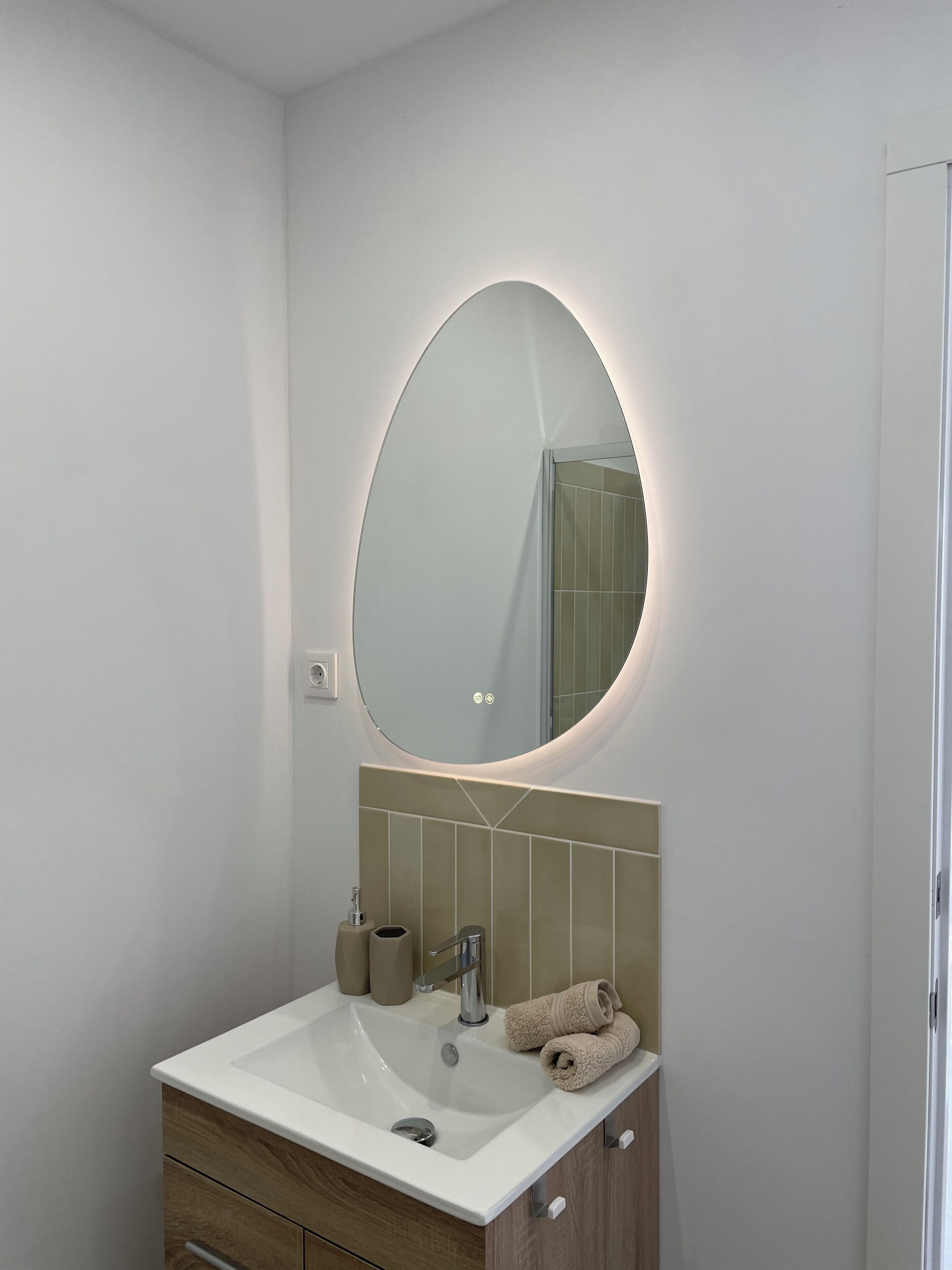 A modern bathroom with a white sink, a mirror with a circular light, beige tiles, and a towel on the counter.