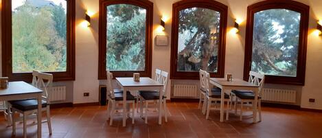 A dining area with four tables and chairs, large arched windows with views of trees, and wall-mounted lighting.