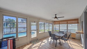 Ein geräumiges Zimmer mit großem Fenster zum Schwimmbad, Deckenventilator und Essbereich mit Stühlen und Tisch.